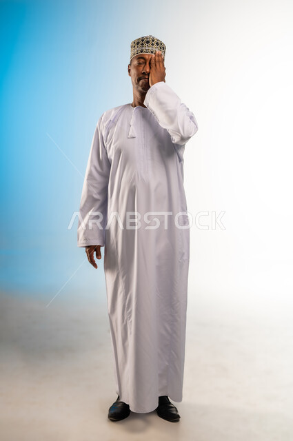 التهاب واحمرار العيون والحاجة لزيارة الطبيب، وضع اليد على العين وتغطيها، ضعف النظر وتشوش الرؤية، التعرض لأذية في الشبكية، بورتريه لرجل عربي خليجي عماني يرتدي الدشداشة والكمة يعاني من مشاكل صحية في عينه، الجسم بالطول الكامل، خلفية ملونة