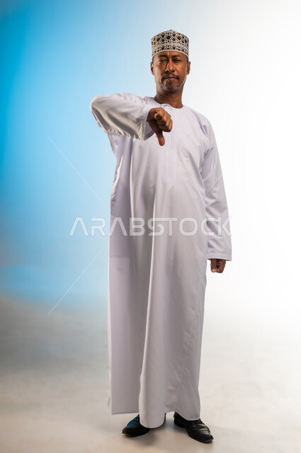 مفهوم الرفض وعدم القبول، بورتريه لرجل عربي خليجي عماني يرتدي الدشداشة و الكمه يشير بإصبع الابهام للاسفل بإيماءات عدم الاعجاب والتقييم السلبي، الشعور بالانزعاج من شيء ما، صورة الجسم بالطول الكامل, خلفية ملونة