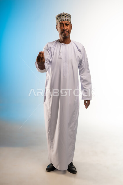 مفهوم الصبر والتريث و التأني, التهديد و التوعد لشيء ما، بورتريه لرجل عربي خليجي عماني يرتدي الدشداشة و الكمه ينظر الى الكاميرا يرفع يده نحو الأعلى بإيماءات طلب الإنتظار, إمهال بعض الوقت لفعل شيء، صورة الجسم بالطول الكامل, خلفية بيضاء
