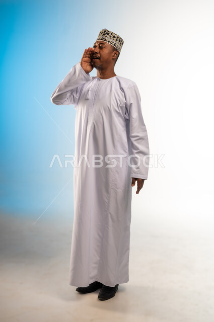 النداء والكلام والتحدث بصوت مرتفع، بورتريه لرجل عربي خليجي عماني مبتسم يرتدي الدشداشة و الكمه يضع يده على فمه بإيماءات المناداة لشخص ما، دردشة ومحادثات وتواصل اجتماعي، صورة الجسم بالطول الكامل, خلفية ملونة