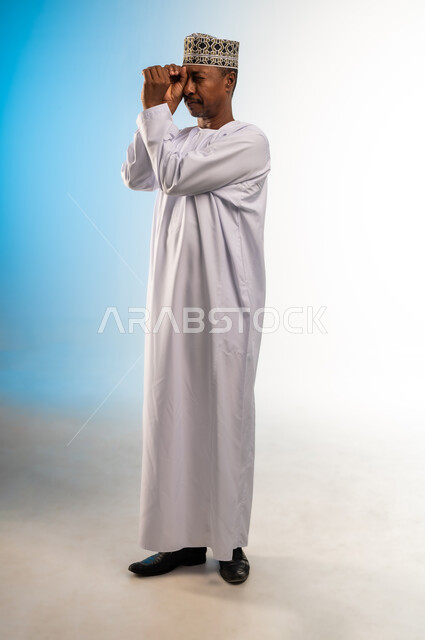 مفهوم التخطيط و الرؤية المستقبلية، بورتريه لرجل عربي خليجي عماني يرتدي الدشداشة و الكمه يضع يده على عينيه بشكل عدسة بإيماءات البحث والاستكشاف، صورة الجسم بالطول الكامل, خلفية ملونة
