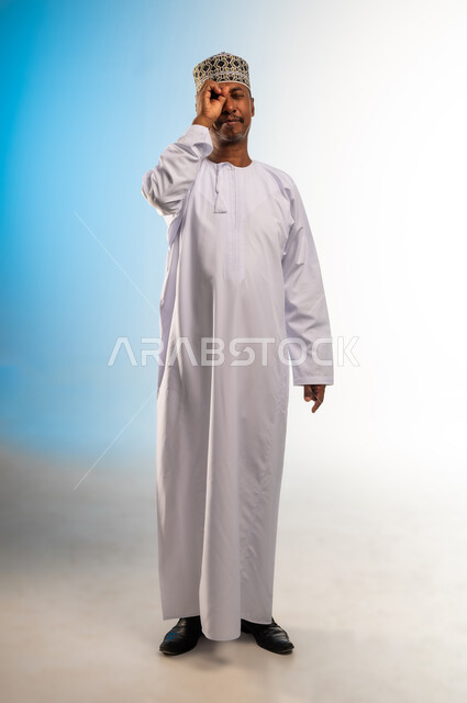 التخطيط و الرؤية المستقبلية، بورتريه لرجل عربي خليجي عماني يرتدي الدشداشة و الكمه يضع يده على عينيه بشكل عدسة بإيماءات البحث والاستكشاف، صورة الجسم بالطول الكامل, خلفية ملونة