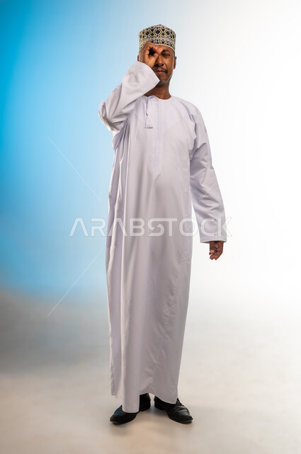 مفهوم التخطيط و الرؤية المستقبلية، بورتريه لرجل عربي خليجي عماني يرتدي الدشداشة و الكمه يضع يده على عينيه بشكل عدسة بإيماءات البحث والاستكشاف، صورة الجسم بالطول الكامل, خلفية ملونة