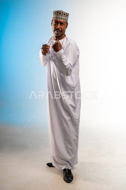 احتراف رياضة الملاكمة، إغلاق اليدين والاستعداد للهجوم والمواجهة، محاولة الدفاع عن النفس، مفهوم القوة والرجولة، بورتريه لرجل عربي خليجي عماني يرتدي الدشداشة والكمة ينظر للكاميرا بإيماءات الجدية والغضب، الجسم بالطول الكامل، خلفية ملونة