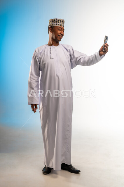 Making a video call, using a modern and advanced technical device, browsing applications and social networking sites on the Internet, chatting, conversations and getting to know each other, portrait of an Arab Gulf Omani man wearing a dishdasha and a cloak using a mobile phone, full body, colored background