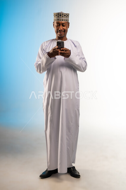 Using a modern technical device, integrating technology into daily life, browsing applications and social networking sites on the Internet, chatting, conversations and getting to know each other, portrait of an Arab Gulf Omani man wearing a dishdasha and a kimono using a mobile phone, full body, colored background