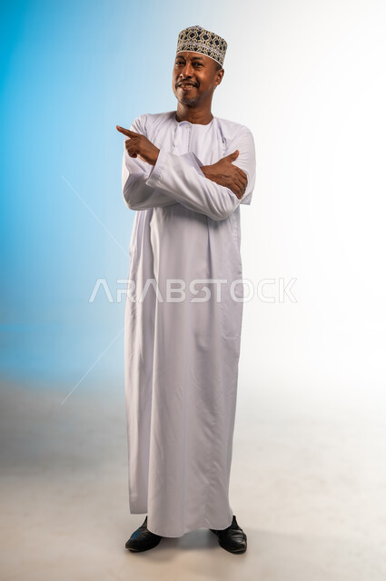تحديد موقع شيء معين، حركة يدين تدل على هذا المكان (هنا)، بورتريه لرجل عربي خليجي عماني مبتسم يرتدي الدشداشة و الكمه  يشير باصبع السبابة، مفهوم الأناقة والاهتمام بالمظهر الخارجي، الجسم بالطول الكامل، خلفيه ملونة
