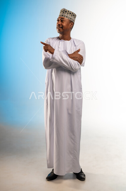 Hand movement indicating this place (here), locating a specific thing, portrait of an Arab Gulf Omani man wearing a dishdasha and a sleeve pointing with his index finger, concept of elegance and interest in external appearance, full-length body, colored background