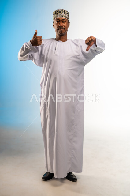 الحيرة والتردد في شيء ما، بورتريه لرجل عربي خليجي عماني مبتسم يرتدي الدشداشة و الكمه ينظر الى الكاميرا يرفع اصبع الابهام للاعلى والاسفل بإيماءات عدم الموافقه والموافقه عدم الإعجاب والإعجاب, مفهوم الرفض والقبول، صورة الجسم بالطول الكامل, خلفية ملونة
