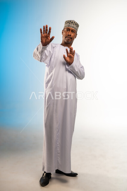 القلق والاحتياط والابتعاد عن شيء ما, حماية النفس من ضرر خارجي,  بورتريه لرجل  عربي خليجي عماني يرتدي الدشداشة و الكمه يرفع يديه للاعلى بإيماءات التوقف وعدم الاقتراب، مفهوم الحذر وترك مسافة، الجسم بالطول الكامل، خلفية ملونة