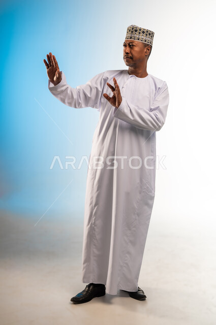 حماية النفس من ضرر خارجي, القلق والاحتياط والابتعاد عن شيء ما, بورتريه لرجل  عربي خليجي عماني يرتدي الدشداشة و الكمه يرفع يديه للاعلى بإيماءات التوقف وعدم الاقتراب، مفهوم الحذر وترك مسافة، الجسم بالطول الكامل، خلفية ملونة