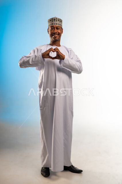 Looking at the camera with gestures of pleasure and happiness, portrait of an Arab Gulf Omani man wearing a dishdasha and a kummah raising his hands in the shape of a heart with expressions of love and affection, full body photo, colored background