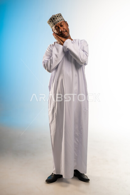 الشعور بالتعب و الحاجه الى الراحه, الإحساس بالكسل والخمول، بورتريه مقرب لرجل عربي خليجي عماني يرتدي الدشداشة و الكمه يضع يديه تحت رأسه يغمض عينيه بإيماءات النوم، تعابير النعاس والإرهاق، صورة الجسم بالطول الكامل، خلفية ملونة