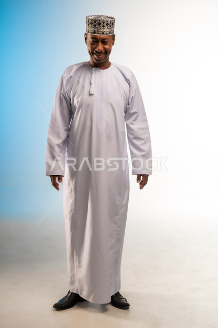 مفهوم الرجولة والثقة بالنفس، التقاط الصور الشخصية للوثائق الرسمية، بورتريه لرجل عربي خليجي عماني مبتسم يرتدي الدشداشة و الكمه ينظر الى الأسفل بإيماءات السرور والسعادة، الاهتمام بالأناقة والمظهر الخارجي، صورة الجسم بالطول الكامل، خلفية ملونة