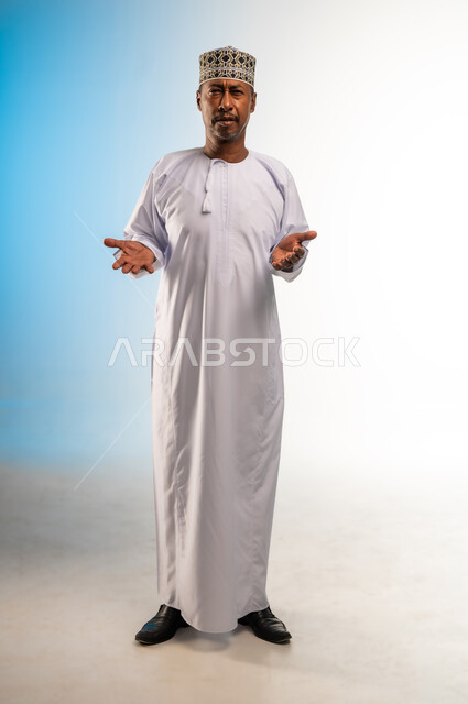 النظر الى الكاميرا بتعابير التردد وعدم الثقة بخصوص أمر معين، بورتريه لرجل عربي خليجي عماني يرتدي الدشداشة و الكمه يرفع كفيه للاعلى بإيماءات عدم التأكد، الشعور بالحيرة وعدم المعرفة، صورة الجسم بالطول الكامل, خلفية ملونة