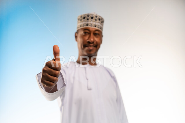 الشعور بالقبول والرضا، النظر الى الكاميرا بتعابير السعادة، بورتريه مقرب لرجل عربي خليجي عماني مبتسم يرتدي الدشداشة و الكمه يرفع اصبع الابهام للاعلى بإيماءات الاعجاب والسرور، علامة الجودة والامتياز، خلفية ملونة
