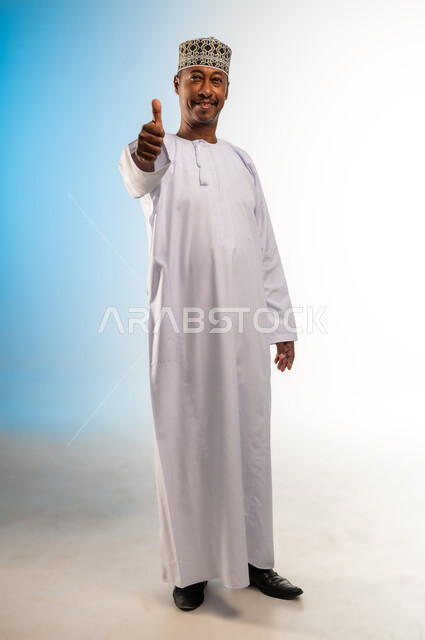 النظر الى الكاميرا بتعابير السعادة، الشعور بالقبول والرضا، بورتريه لرجل عربي خليجي عماني يرتدي الدشداشة و الكمه يرفع اصبع الابهام للاعلى بإيماءات الاعجاب والسرور، علامة الجودة والامتياز، صورة الجسم بالطول الكامل، خلفية ملونة