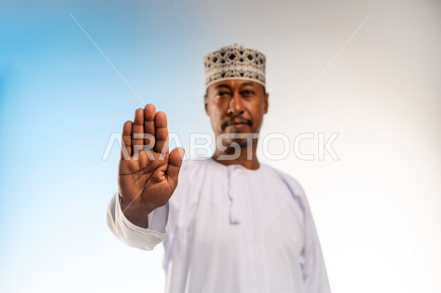 Expressions of refusal and abstention, order to stop doing something, close-up portrait of an Arab Gulf Omani man wearing a dishdasha and a cloak raising his hand up in a gesture of prohibition, feeling annoyed and angry, colored background