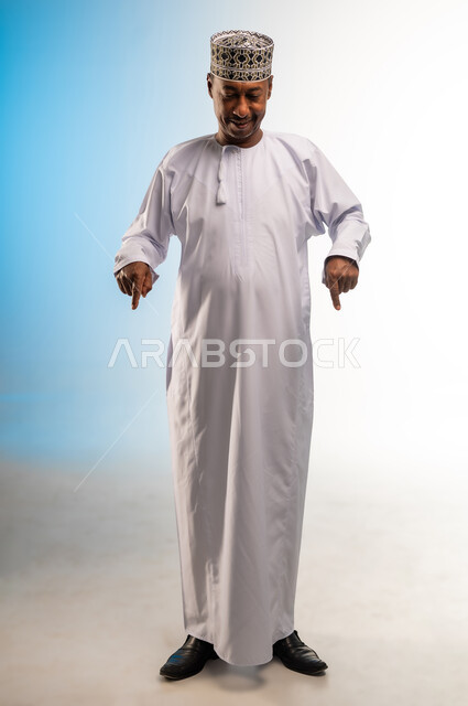 تحديد موقع معين، تأكيد حدوث شيء هذه اللحظة, حركات تدل على هذا المكان (هنا)، بورتريه لرجل عربي خليجي عماني يرتدي الدشداشة و الكمه يقف باستقامة يشير بإصابع السبابة نحو الأسفل, النظر إلى شيء ما بايماءات السرور، صورة الجسم بالطول الكامل, خلفية ملونة