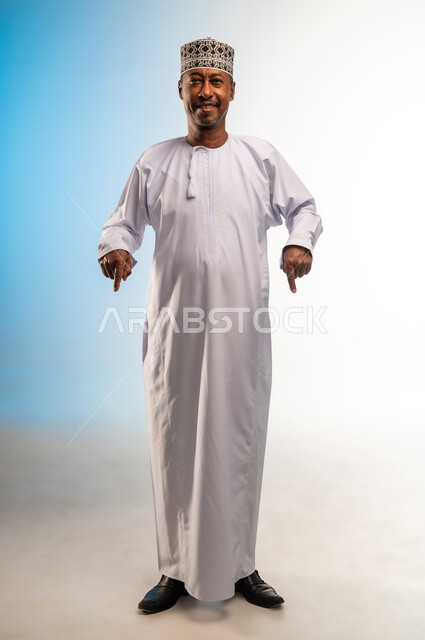 تأكيد حدوث شيء هذه اللحظة, حركات تدل على هذا المكان (هنا)، تحديد موقع معين، بورتريه لرجل عربي خليجي عماني يرتدي الدشداشة و الكمه يقف باستقامة يشير بإصابع السبابة نحو الأسفل, النظر إلى الكاميرا بايماءات السرور، صورة الجسم بالطول الكامل, خلفية ملونة
