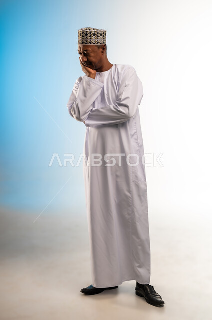 Feeling bored and upset, contemplating and concentrating to find a solution to a problem, solving problems and making decisions, portrait of an Arab Gulf Omani man wearing a dishdasha and a sleeve, putting his hand on his face with gestures of thinking, confusion and annoyance, full body image, colored background