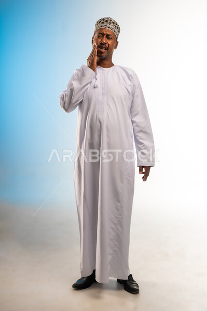 تسوس الأضراس وإلتهاب اللثه، طلب العلاج و المساعده، بورتريه لرجل عربي خليجي عماني يرتدي الدشداشة و الكمه يضع يديه على خده ويغمض عينيه يشعر بألم الأسنان، الحاجه لزيارة الطبيب، صورة الجسم بالطول الكامل, خلفية ملونة
