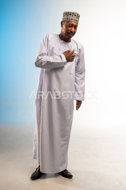 اضطرابات وأمراض قلبية، الإحساس بذبحة صدرية، إيماءات التعب والإرهاق والضغوطات النفسية، بورتريه لرجل عربي عماني خليجي يرتدي الدشداشة و الكمه يضع يده على قلبه يشعر بحالة سيئة من الوخز والألم في قلبه، صوره الجسم بالطول الكامل، خلفية ملونة