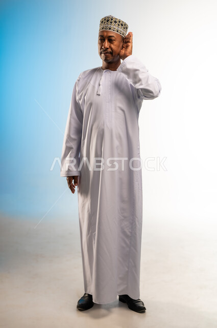 محاولة التجسس والتنصت, حسن الإستماع و التركيز على الكلام, سماع الأسرار والمحادثات، بورتريه لرجل عربي عماني خليجي يرتدي الكمه و الدشداشة يضع يده خلف اذنه, صورة الجسم بالطول الكامل، خلفية ملونة