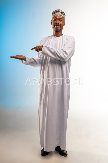 Pointing with index finger to something specific, standing and looking at camera with gestures of pleasure, portrait of an Arab Omani Gulf man wearing a turban and a dishdasha holding something in his hand, lifting and carrying something specific, full body, colored background