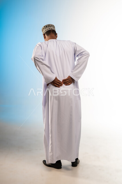 الحاجة إلى العلاج وزيارة الطبيب، التعب والإرهاق والإحساس بالمرض، آلام العامود الفقري والظهر، بورتريه من الخلف لرجل عربي خليجي عماني يرتدي الكمه و الدشداشة يضع يده على ظهره بتعابير الانحناء، الشعور بالوجع والألم، صورة الجسم بالكامل، خلفية ملونة
