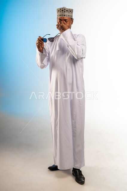 تذكر شيء محدد بشكل مفاجئ, ايماءات الالم من أشعه الشمس, بورتريه لرجل عربي خليجي عماني يرتدي الكمه و الدشداشة يحمل النظارات الشمسية و يضع يده على وجهه ويضغط على عينيه بتعابير الوجع الشديد, صورة الجسم بالطول الكامل, خلفية ملونة