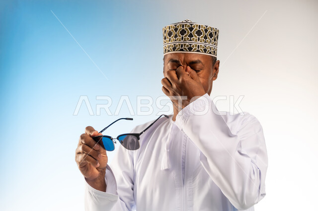 ايماءات الالم من أشعه الشمس, تذكر شيء محدد بشكل مفاجئ, بورتريه مقرب لرجل عربي خليجي عماني يرتدي الكمه و الدشداشة يحمل النظارات الشمسية و يضع يده على وجهه ويضغط على عينيه بتعابير الوجع الشديد, خلفية ملونة