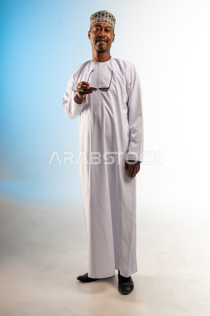 النظر الى الكاميرا بايماءات السرور, الاهتمام بالمظهر الخارجي, بورتريه لرجل عربي خليجي عماني يرتدي الكمه و الدشداشة يحمل بيده النظارات الشمسية, مفهوم الاناقة  و الرجولة, صورة الجسم بالطول الكامل, خلفية ملونة