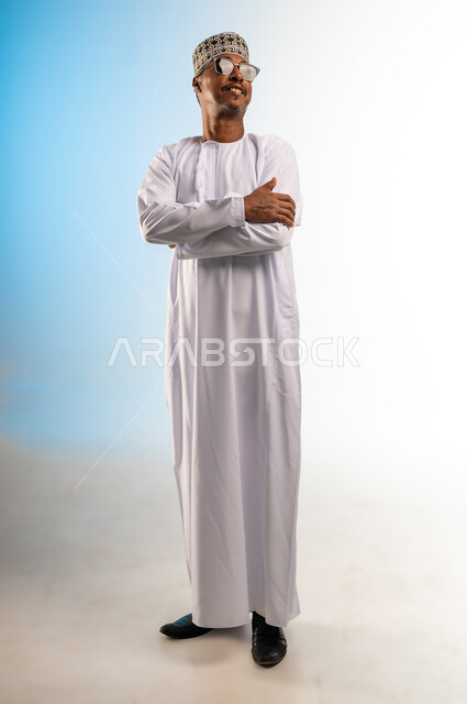 الاهتمام بالمظهر الخارجي, بورتريه لرجل عربي خليجي عماني يرتدي الكمه و الدشداشة و النظارات الشمسية يقف بتعابير أيدي متقاطعه, النظر الى شيء ما بايماءات السرور, مفهوم الاناقة  و الرجولة, صورة الجسم بالطول الكامل, خلفية ملونة