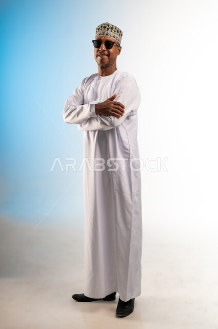 The concept of masculinity and self-confidence, interest in elegance and appearance, portrait of a smiling Arab Gulf Omani man wearing a dishdasha, a sleeve and sunglasses, standing with crossed hands looking at the camera with gestures of pleasure, full-length body image, colored background