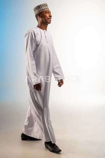 مفهوم الرجولة والثقة بالنفس، الشعور بالفرح والسرور، الاهتمام بالأناقة والمظهر الخارجي، بورتريه من الجانب لرجل عربي خليجي عماني يرتدي الدشداشة و الكمه يمشي الى الامام ينظر الى شيء ما، إيماءات وتعابير الذهاب لمكان ما، صورة الجسم بالطول الكامل، خلفية ملونة