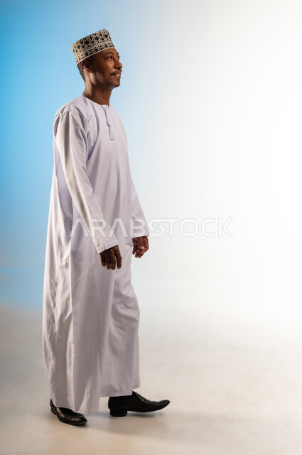الشعور بالفرح والسرور، الاهتمام بالأناقة والمظهر الخارجي، مفهوم الرجولة والثقة بالنفس، بورتريه من الجانب لرجل عربي خليجي عماني يرتدي الدشداشة و الكمه يمشي الى الامام ينظر الى شيء ما، إيماءات وتعابير الذهاب لمكان ما، صورة الجسم بالطول الكامل، خلفية ملونة