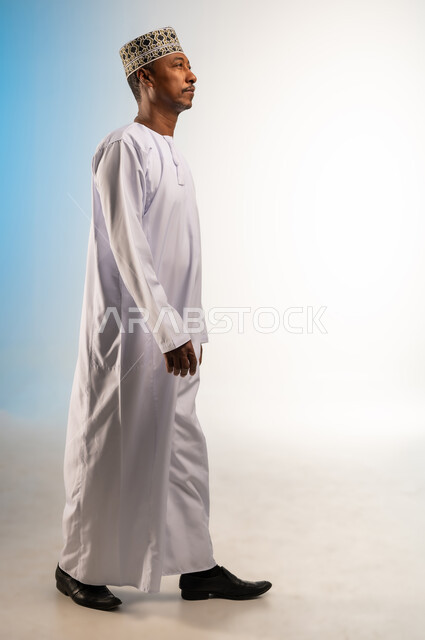 الاهتمام بالأناقة والمظهر الخارجي، مفهوم الرجولة والثقة بالنفس، بورتريه من الجانب لرجل عربي خليجي عماني يرتدي الدشداشة و الكمه يمشي الى الامام ينظر الى شيء ما، إيماءات وتعابير الذهاب لمكان ما، صورة الجسم بالطول الكامل، خلفية ملونة