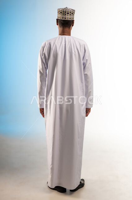 مفهوم الرجولة والثقة بالنفس، الاهتمام بالأناقة والمظهر الخارجي، بورتريه من الخلف لرجل عربي خليجي عماني يرتدي الدشداشة و الكمه يقف باستقامة ينظر الى شيء ما، صورة الجسم بالطول الكامل، خلفية ملونة