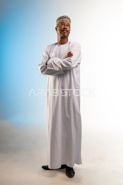 مفهوم الرجولة والثقة بالنفس، التقاط الصور الشخصية للوثائق الرسمية، بورتريه لرجل عربي خليجي عماني مبتسم يرتدي الدشداشة و الكمه يقف بأيدي متقاطعة ينظر الى شيء ما بالأعلى، الاهتمام بالأناقة والمظهر الخارجي، صورة الجسم بالطول الكامل، خلفية ملونة