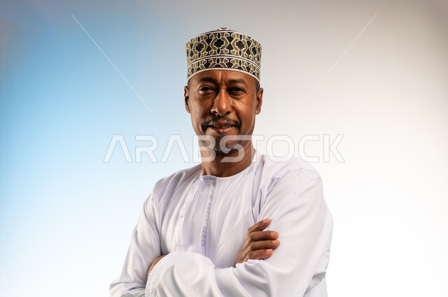 إيماءات التفكير وشرود الذهن، مفهوم الرجولة والثقة بالنفس، بورتريه مقرب لرجل عربي خليجي عماني مبتسم يرتدي الدشداشة و الكمه يقف بأيدي متقاطعة ينظر الى الكاميرا بتعابير السرور، الاهتمام بالأناقة والمظهر الخارجي، خلفية ملونة