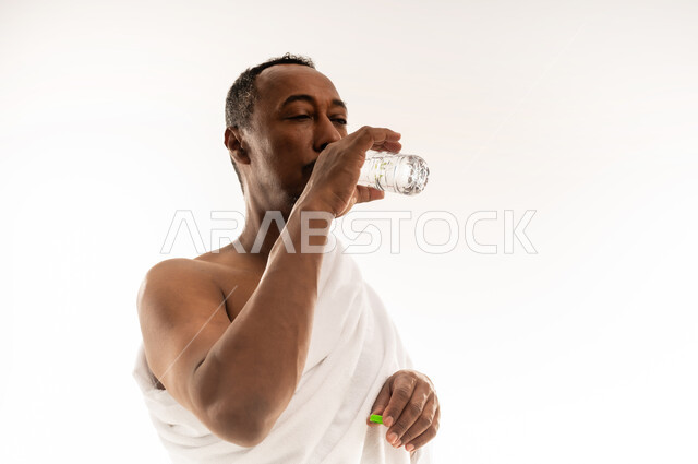 حاج سعودي يشعر بالعطش والجفاف، المحافظة على سلامة الجهاز المناعي للجسم، المداومة على شرب المياه، بورتريه مقرب لرجل عربي عماني خليجي يرتدي لباس الاحرام يحمل بيده القارورة يشرب ماء زمزم بإيماءات الارتواء، خلفية بيضاء
