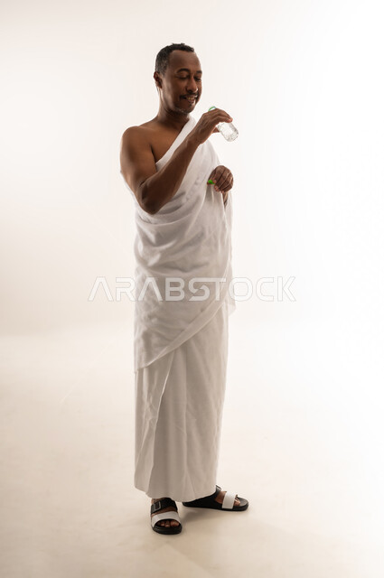المداومة على شرب المياه، المحافظة على سلامة الجهاز المناعي للجسم، حاج سعودي يشعر بالعطش والجفاف، بورتريه لرجل عربي عماني خليجي يرتدي لباس الاحرام يحمل بيده قارورة من ماء زمزم بإيماءات الفرح والسرور، صورة الجسم بالطول الكامل، خلفية بيضاء