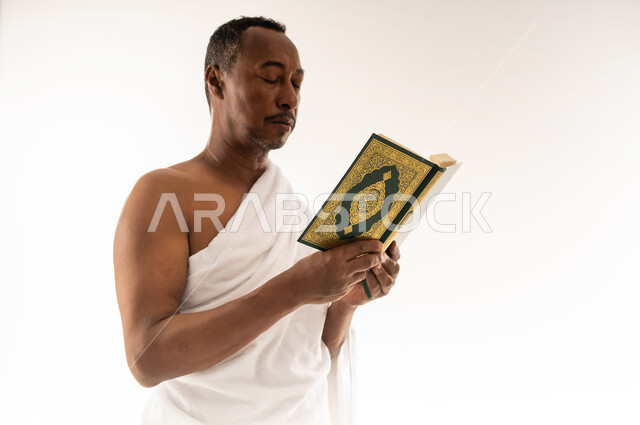 مفهوم العبادة و التقرب الى الله, حاج سعودي يقوم بتلاوة المصحف الشريف، الشعور بالسكينة والراحة، اداء مناسك الحج والعمرة، بورتريه مقرب لرجل عربي عماني خليجي يرتدي لباس الاحرام يحمل بيده القرآن الكريم، خلفية بيضاء