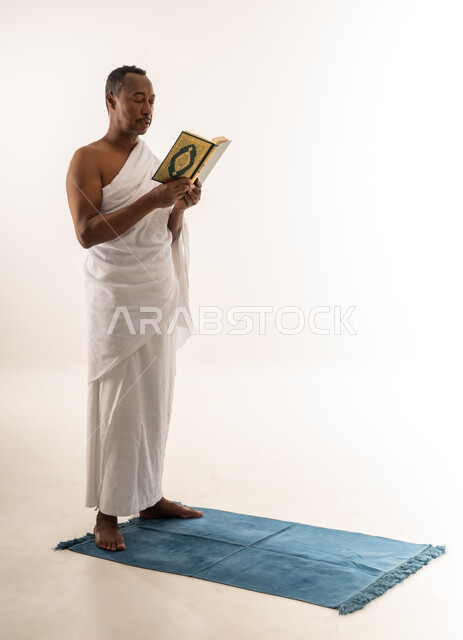 حاج سعودي يقوم بتلاوة المصحف الشريف، الشعور بالسكينة والراحة، مفهوم العبادة و التقرب الى الله, اداء مناسك الحج والعمرة، بورتريه لرجل عربي عماني خليجي يرتدي لباس الاحرام يقف على سجادة الصلاة يحمل بيده القرآن الكريم، صورة الجسم بالطول الكامل، خلفية بيضاء