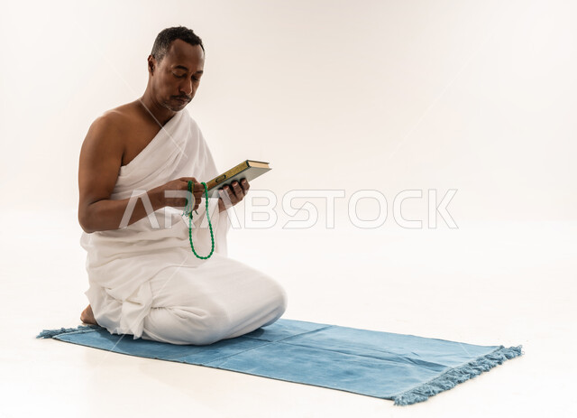 اداء مناسك الحج والعمرة، رجل سعودي يقرأ المصحف الشريف، مفهوم العبادة و التقرب الى رب العالمين, بورتريه لحاج عربي عماني خليجي يرتدي لباس الاحرام يجلس على سجادة الصلاة و يحمل بيده السبحة و القرآن الكريم، خلفية بيضاء
