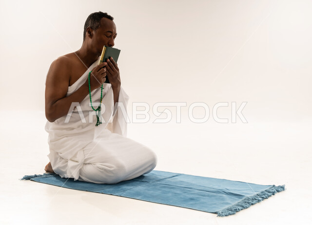 مفهوم العبادة و التقرب الى الله, رجل سعودي يقراءة المصحف الشريف، بورتريه لحاج عربي عماني خليجي يرتدي لباس الاحرام يجلس على سجادة الصلاة و يحمل بيده السبحة و القرآن الكريم يقوم بتقبيله، اداء مناسك الحج والعمرة، خلفية بيضاء