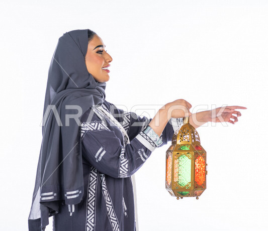 بورتريه لمرأة عربية خليجية سعودية ، تحمل بيدها فانوس ملون ، تشير بيدها نحو شيء محدد أو شخص ما، بإيماءات يد مختلفة، النظر بالإتجاه المحدد ، منظر جانبي لشابة سعودية محجبة ترتدي العباءة الخليجية، خلفية بيضاء، العبادة والتقرب إلى الله ، فانوس رمضان