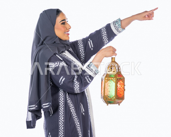 بورتريه لمرأة عربية خليجية سعودية ، تحمل بيدها فانوس ملون ، تشير بإصبع السبابة لأعلى ، بإيماءات وجه ويدين مختلفة ، منظر جانبي لشابة سعودية مسلمة محجبة ترتدي العباءة الخليجية ، خلفية بيضاء، العبادة والتقرب إلى الله ، فانوس رمضان