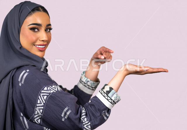 بورتريه لمرأة عربية خليجية سعودية مبتسمة تنظر إلى الكاميرا ، تقوم بإيماءات يد مختلفة ، تشير بإصبعها السبابة على يدها الأخرى ، تحمل عنصر هولوجرامي ، شابة سعودية مسلمة محجبة ترتدي العباءة الخليجية ، خلفية بنفسجية